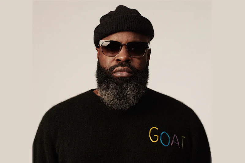 Black Thought in a minimalist studio portrait wearing a black beanie, dark sunglasses, and a black sweater with subtle “GOAT” embroidery, his full beard sharply defined against a neutral backdrop