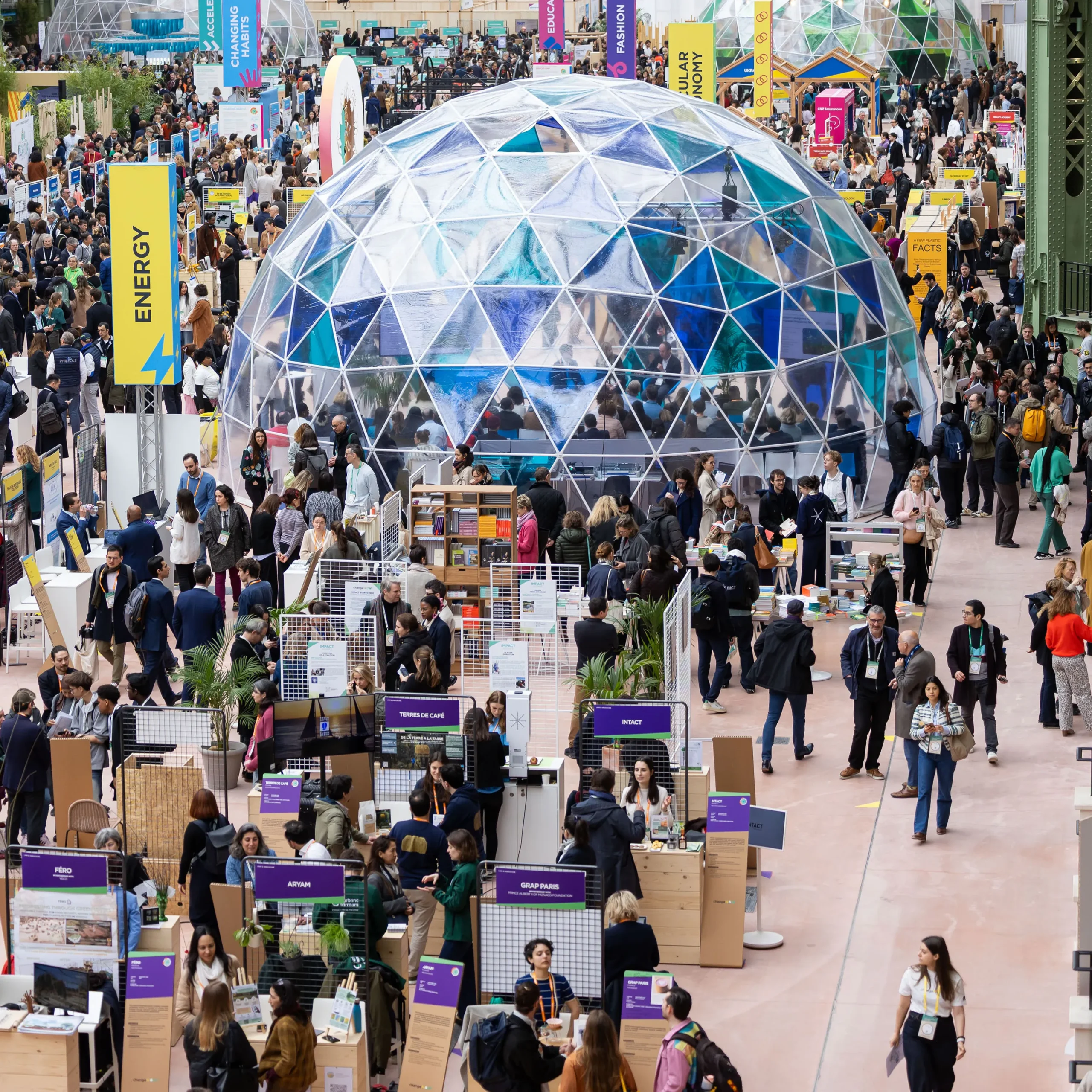 Crowded sustainability conference floor centered around a large transparent geodesic dome, with attendees moving between exhibition booths, climate innovation displays, and vertical banners highlighting themes like energy and circular economy