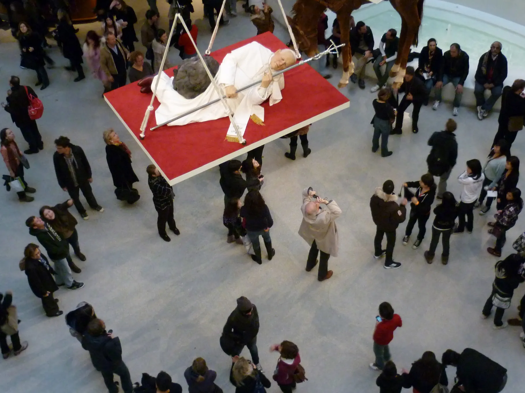 Installation view of Maurizio Cattelan La Nona Ora showing a wax figure of Pope John Paul II struck by a meteor, displayed on a suspended red platform above museum visitors observing the work from below