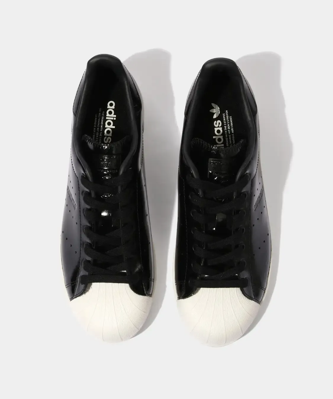 Adidas Superstan in black patent leather shown from the front, featuring glossy uppers, black laces, and the signature white Superstar shell toe contrasting against the sleek Stan Smith-inspired design