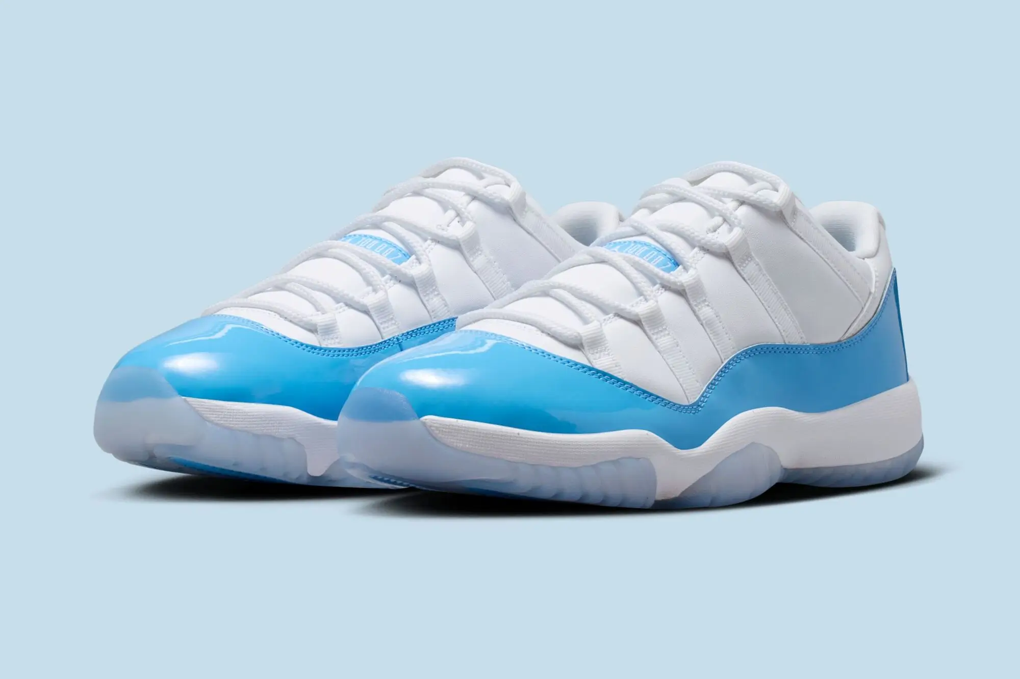 Primary Alt Text (Mid): Air Jordan 11 Low UNC 2026 pair shown in angled perspective with white leather upper, Carolina blue patent leather mudguard, and icy translucent outsole