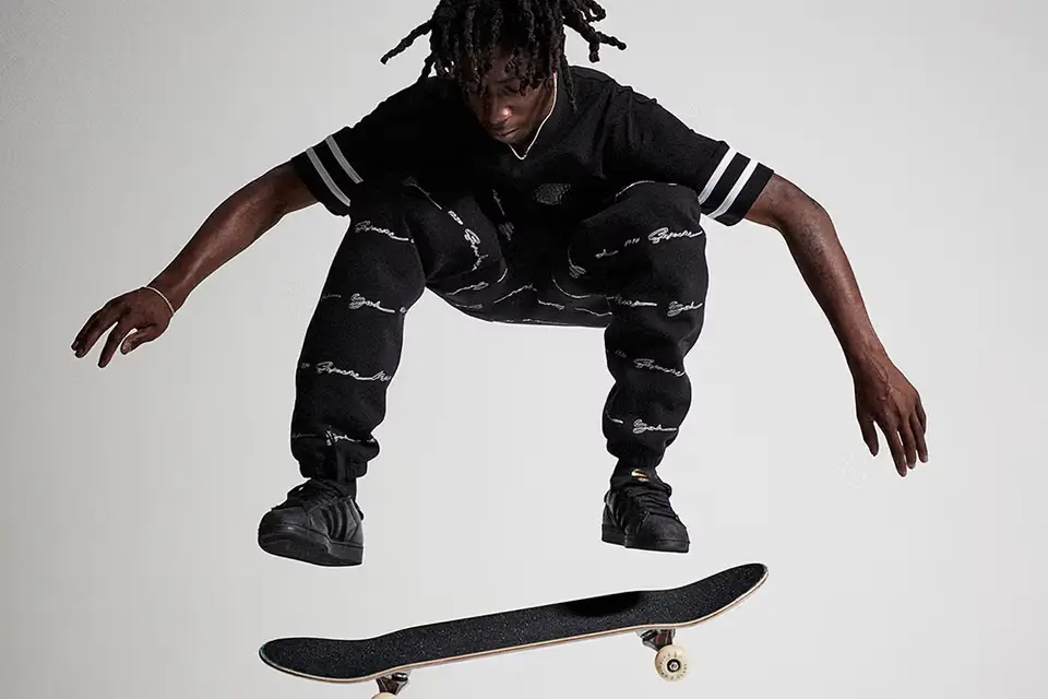 Kader Sylla captured mid-air performing a controlled flip trick against a clean studio backdrop, wearing an all-black outfit with patterned pants and adidas skate shoes. His body remains compact and centered above the rotating board, emphasizing precision, balance, and the restrained technical style that defines his approach