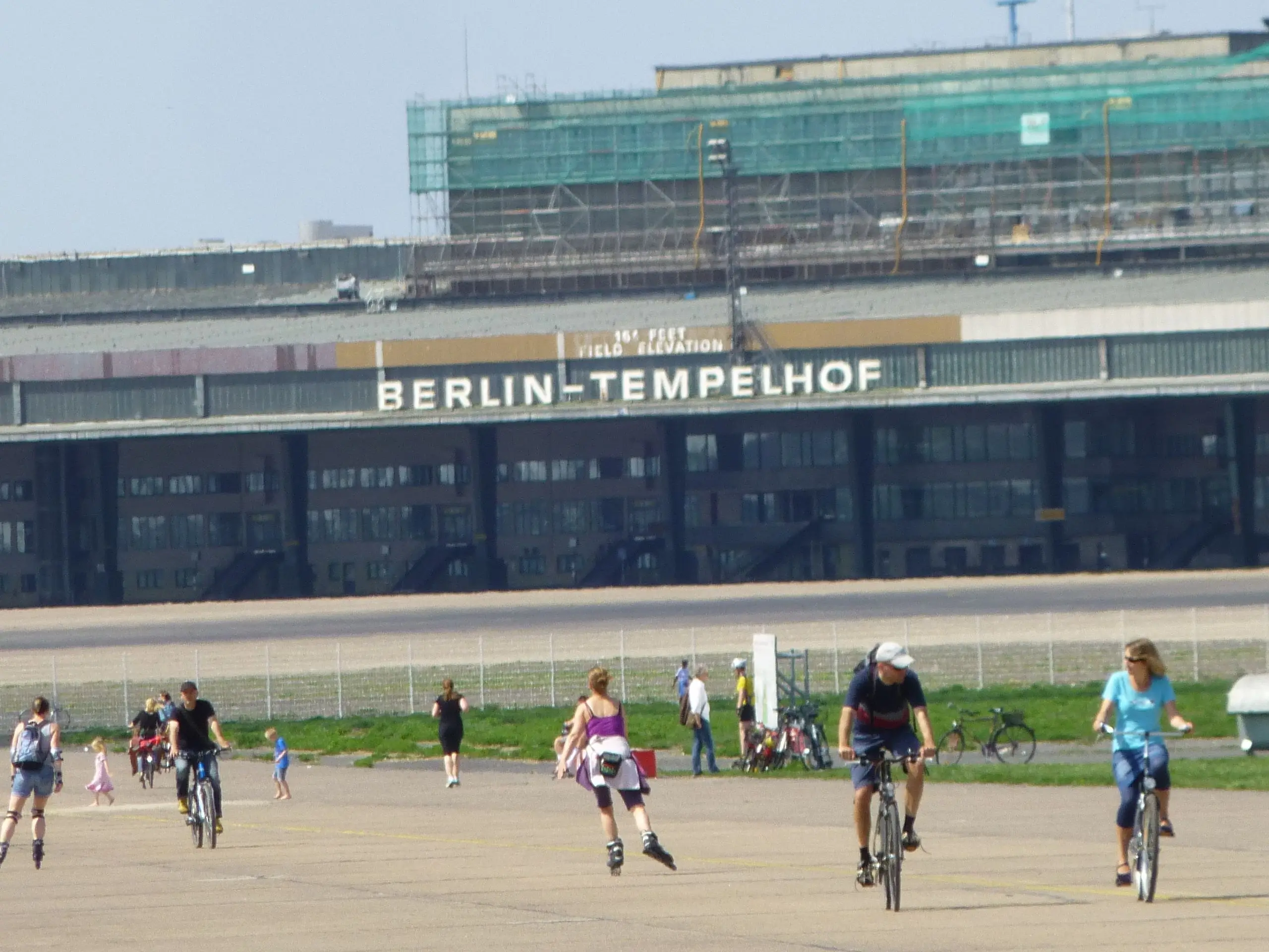 Wide view of Tempelhofer Feld runway in Berlin with people cycling, skating, and walking in front of the historic Berlin-Tempelhof airport building in the background