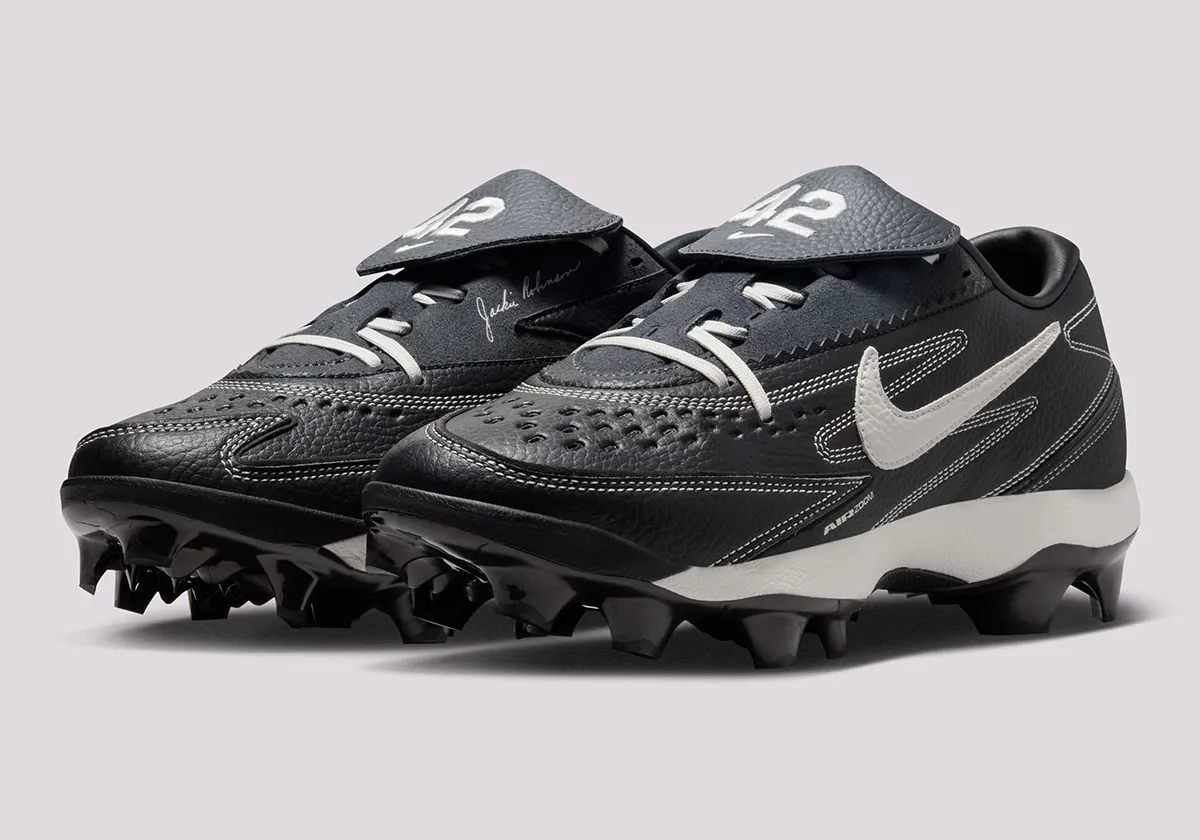 Three-quarter view of the Nike Diamond Standout MCS “Jackie Robinson” cleats featuring a black leather upper with white contrast stitching, perforated toe box, oversized white Swoosh, molded black studs, and tongue flaps marked with #42 above Jackie Robinson’s signature detail