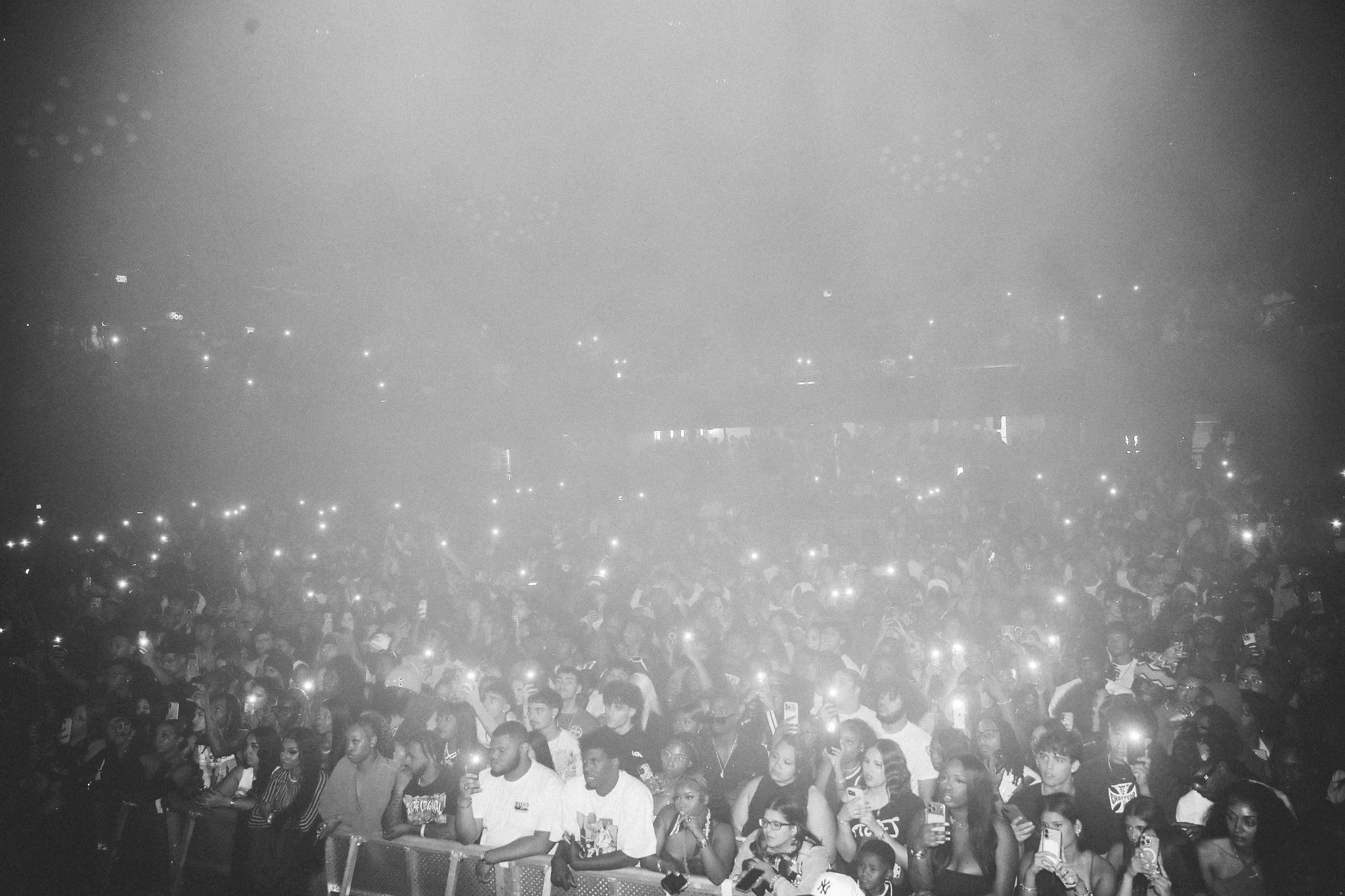 Black-and-white crowd shot at Loe Shimmy’s Broward County birthday concert, with a packed audience holding up phones, glowing lights scattered across the venue, and fans gathered tightly behind the front barrier