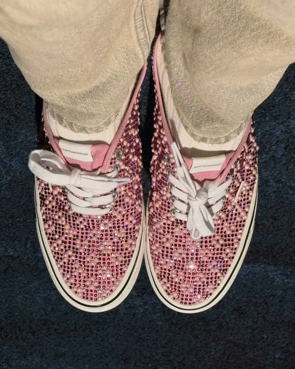 Top-down view of pink embellished Vans Authentic sneakers covered in rhinestones and pearl-like studs, paired with white laces and worn with beige pants against a dark textured surface