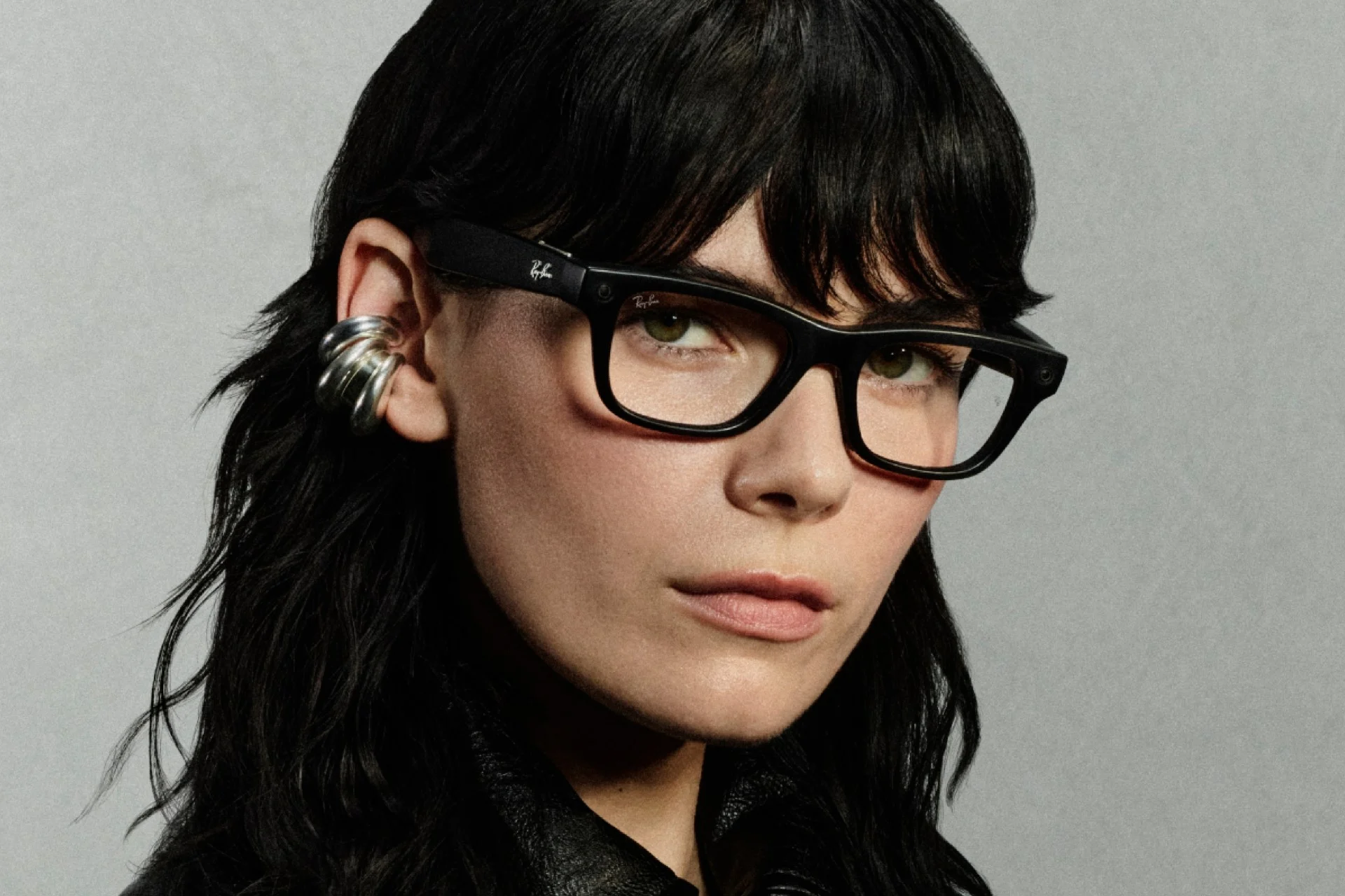 Person wearing black rectangular Meta x Ray-Ban prescription smart glasses with integrated front cameras, styled with a silver ear cuff and dark layered hair against a neutral backdrop