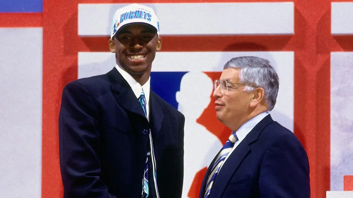 Young Kobe Bryant smiles on stage at the 1996 NBA Draft wearing a Charlotte Hornets cap and dark suit, standing beside NBA Commissioner David Stern in front of the league logo backdrop