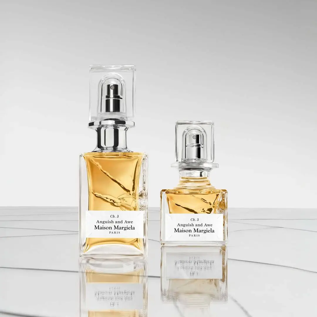 Two Maison Margiela haute parfumerie bottles labeled “Anguish and Awe,” presented in sculptural clear glass with faceted caps and warm amber liquid, set against a minimalist reflective surface emphasizing clarity and refined composition