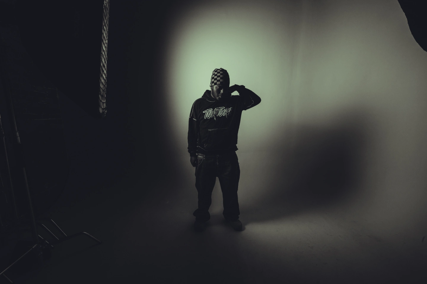 Slayr stands in a dimly lit studio wearing a black hoodie and checkered head covering, captured in a moody, shadow-heavy portrait reflecting his atmospheric rap aesthetic