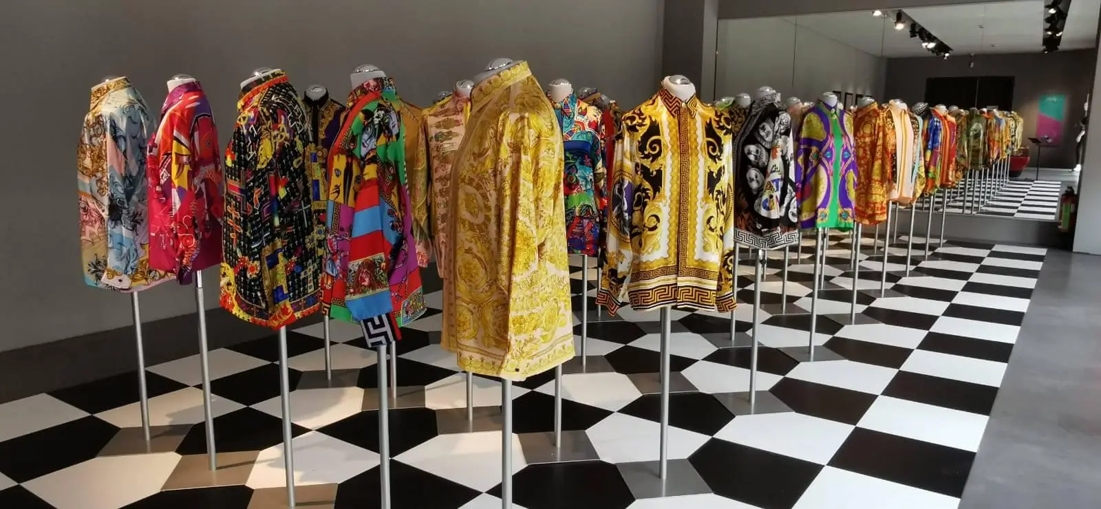 Installation view of Gianni Versace retrospective featuring a long row of mannequins dressed in vibrant baroque-print shirts and garments, arranged in a gallery space with a black-and-white geometric floor, emphasizing Versace’s bold use of color, pattern, and repetition
