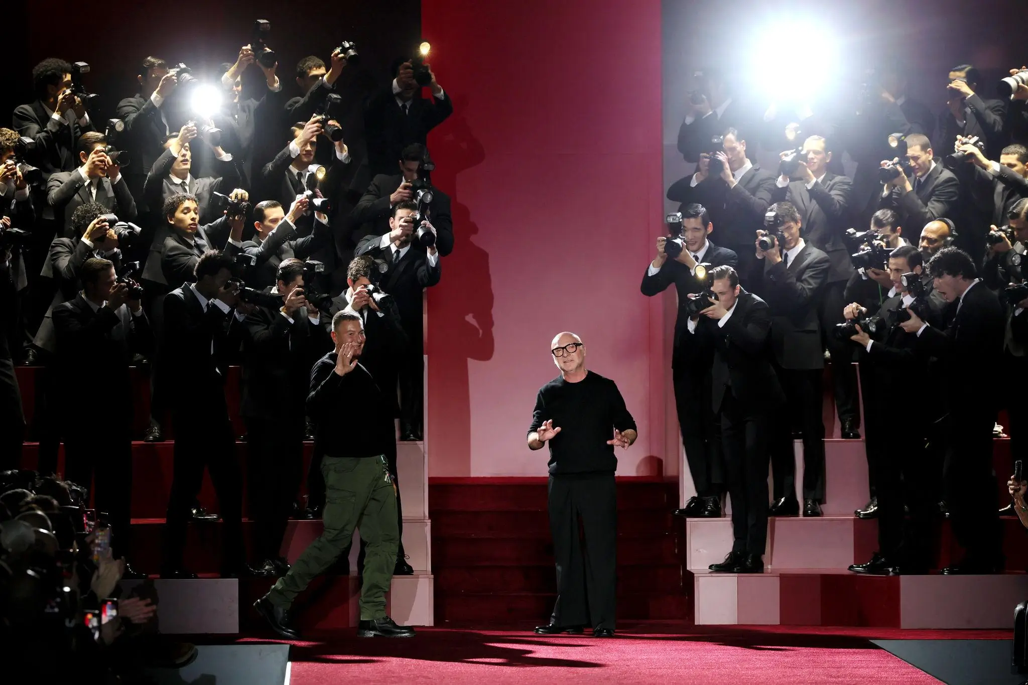 Two designers stand on a red runway stage, surrounded by photographers in black suits whose cameras flash brightly, capturing the moment as the pair acknowledge the crowd in a dramatic, high-fashion show setting