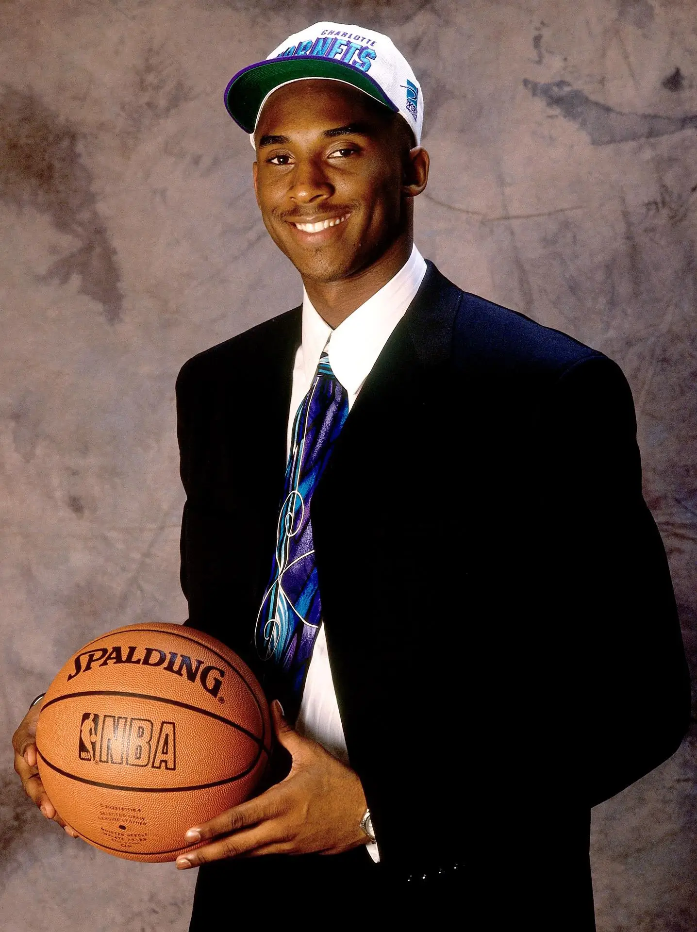 Young Kobe Bryant poses with a basketball at the 1996 NBA Draft, wearing a Charlotte Hornets cap and a dark suit, smiling confidently against a neutral backdrop