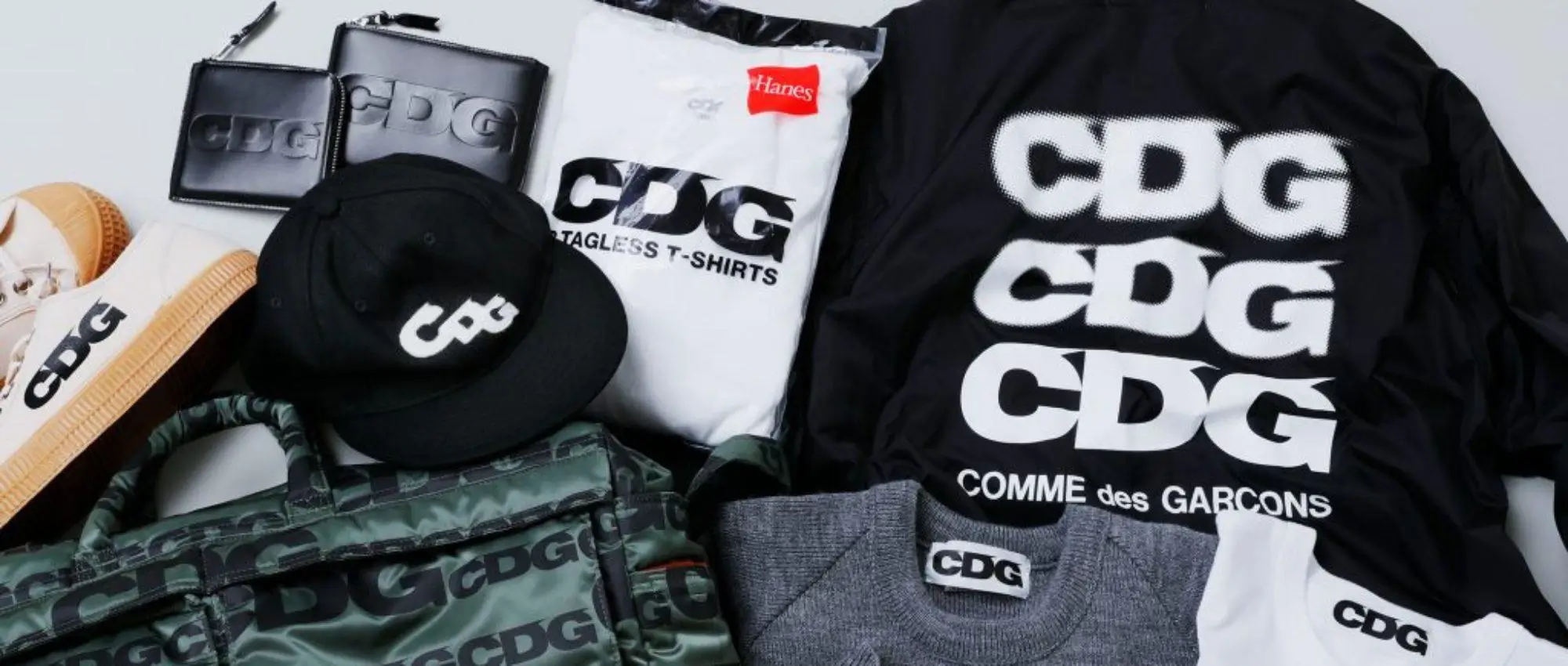 Flat lay of CDG accessories and apparel including logo-heavy coach jacket, cap, wallet, sneakers, bag, and Hanes collaboration T-shirts, emphasizing bold typography branding