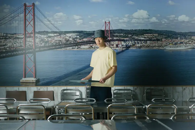 model in a yellow t-shirt and cap stands in a cafeteria setting with empty metal chairs, framed by a large cityscape mural, reflecting wetheknot’s focus on routine environments