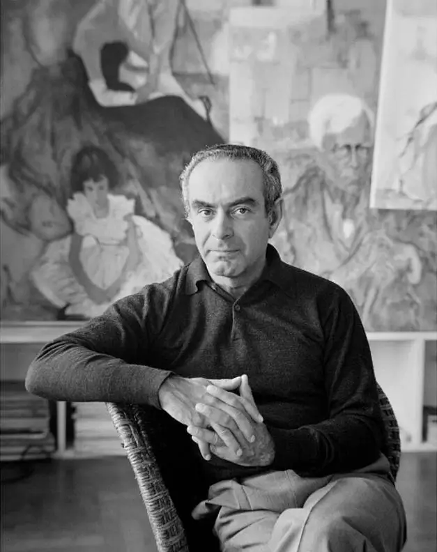 Black-and-white portrait of René Bouché seated with hands clasped, posed before a backdrop of figurative paintings in his studio